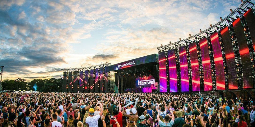 Thousands of festival-goers watch a live outdoor performance as a huge stage with bright vertical LED panels glows at sunset at Creamfields.