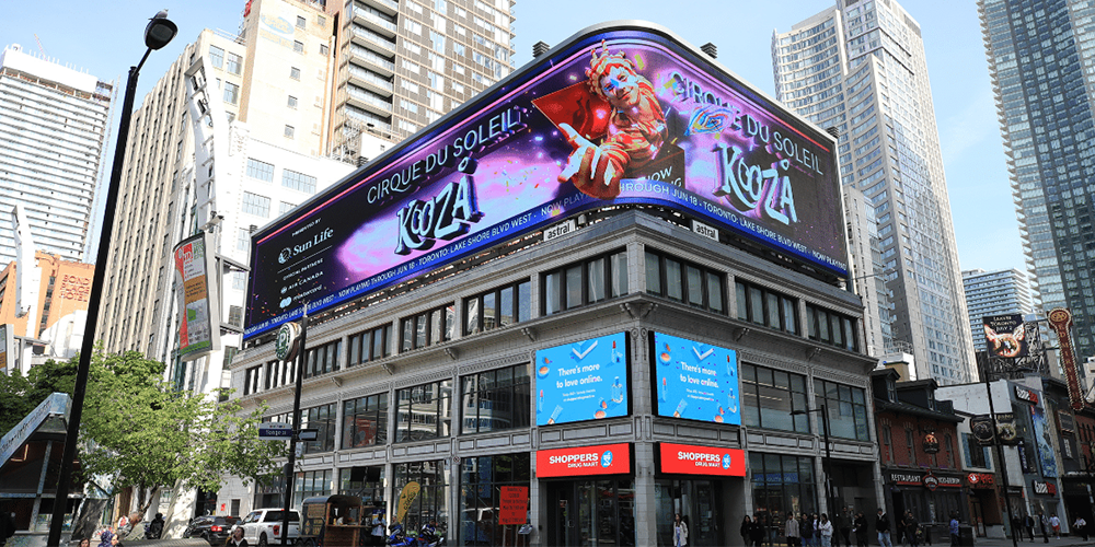 City street scene with a large curved billboard atop a modern building advertising Cirque du Soleil KOZA show in Toronto/Streetscape backdrop of tall buildings.