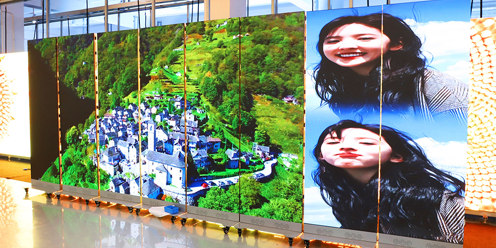 LED poster display-4 - LED Screen Board - LED Screen Board Multi-panel display showing a green hillside village on the left and a portrait of a smiling woman on the right, mounted on wheeled stands, in a bright indoor space.