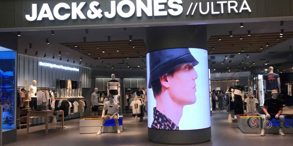 Jack & Jones Ultra storefront with a tall cylindrical screen displaying a man’s side profile, inside a clothing store with mannequins and racks of apparel.