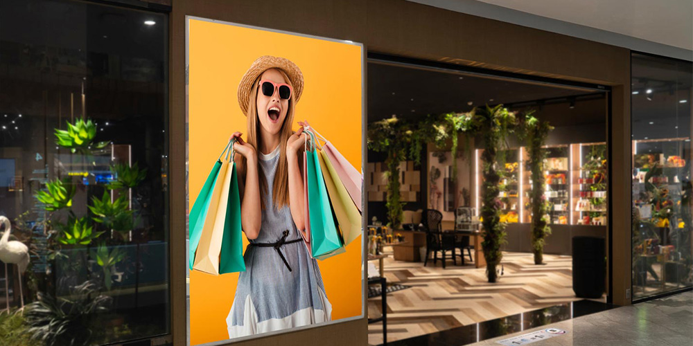 Poster of a smiling woman wearing a hat and sunglasses, holding colorful shopping bags against a yellow background in a mall storefront.