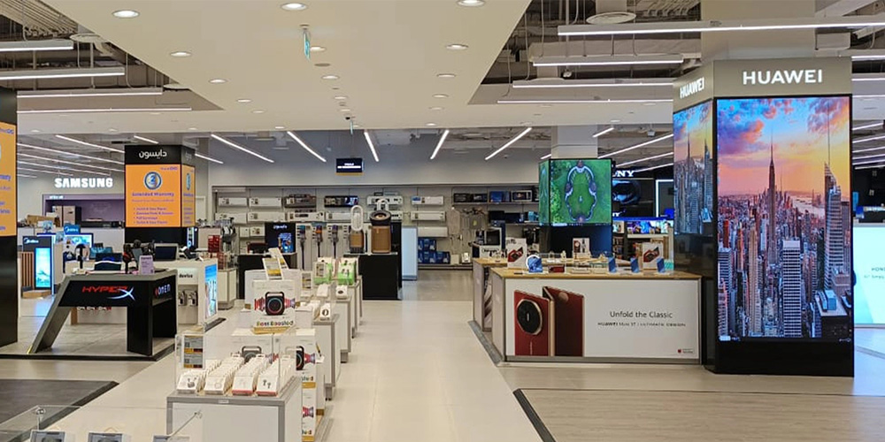 Bright electronics store interior with product counters, shelves, and large Huawei and Samsung displays in a well-lit showroom.