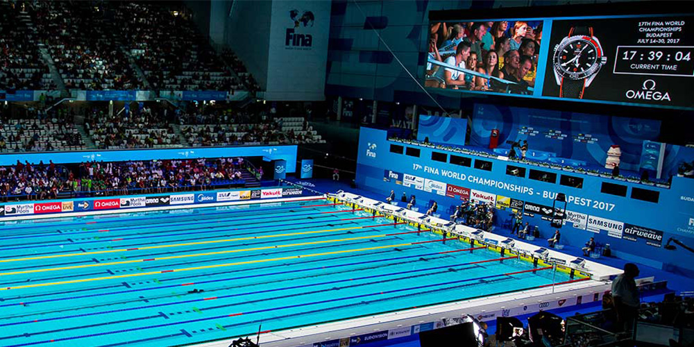 Indoor swimming arena with multiple blue lane pools and spectator seating in the background, large display screens above scoreboard area.