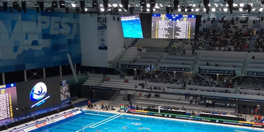 体2 Indoor swimming arena with a blue pool, bleachers, and a large overhead scoreboard showing race details and scores.