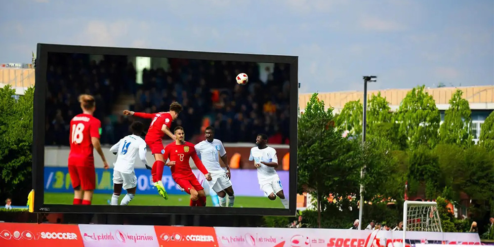 体3 Large outdoor stadium screen showing a soccer match with players in red and white kits near the ball in midair, surrounded by green trees and banners.