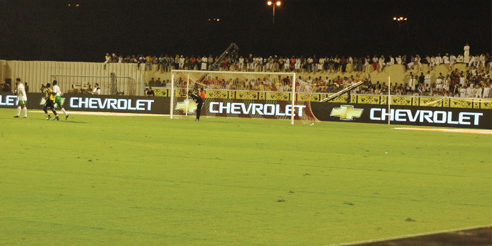 体4 Soccer field at night with players near a goal and bright Chevrolet banners along the sideline; stands full of spectators in the background.