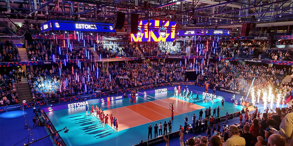 体9 - LED Screen Board - LED Screen Board Packed indoor volleyball arena with a turquoise court; teams stand in a line as fireworks flare on the right and a large cheering crowd watches, Estonia banners visible.