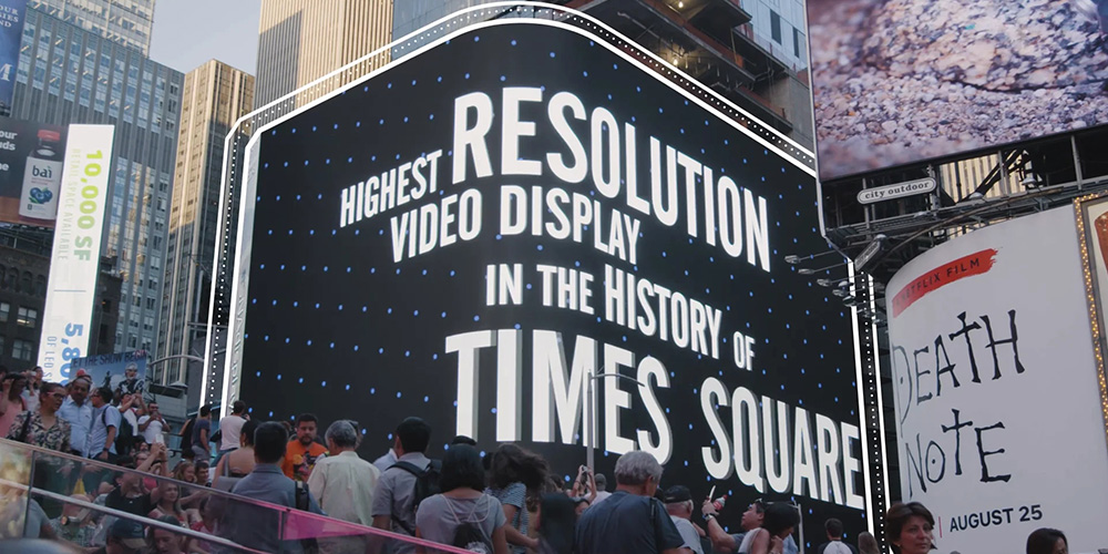 Crowd in Times Square gathered before a giant curved LED display that reads 'Highest Resolution Video Display in the History of Times Square'.