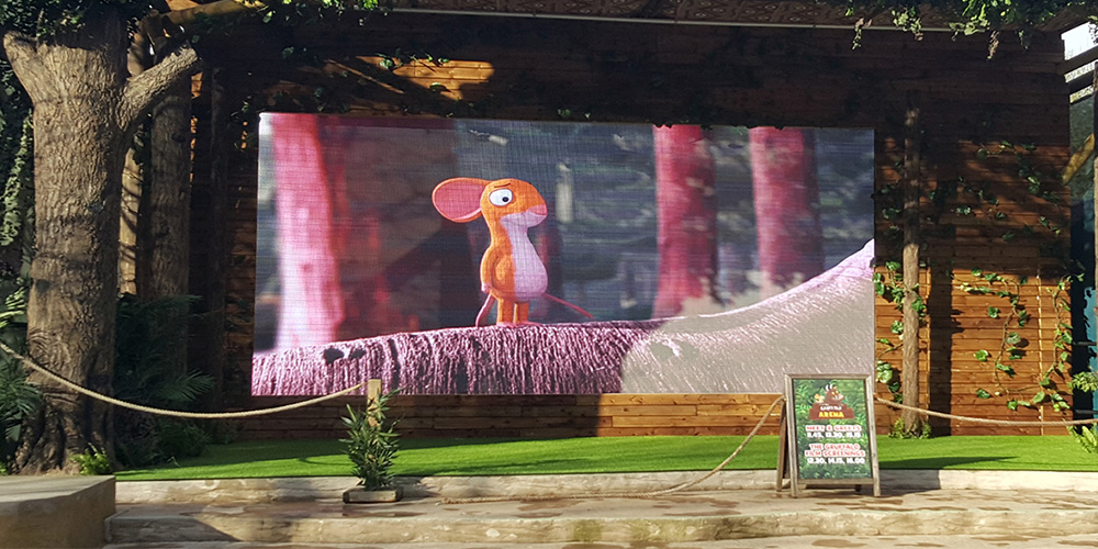 租2 - LED Screen Board - LED Screen Board Outdoor cinema setup on a wooden stage, a large screen showing a cartoon mouse, with greenery and a chalkboard sign nearby.