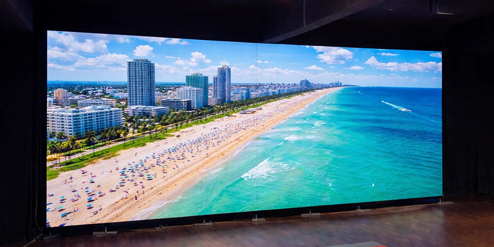 租2y - LED Screen Board - LED Screen Board Aerial view of a sunny beach with turquoise water, a long sandy shore, and high‑rise buildings along the coast with many umbrellas.