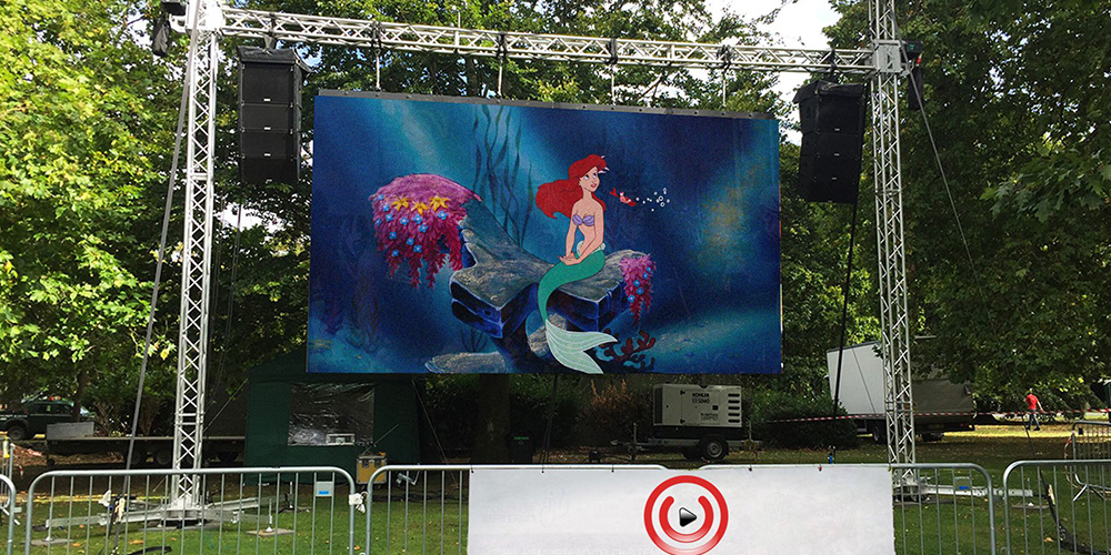 租4 - LED Screen Board - LED Screen Board Outdoor stage with a large screen showing Ariel from The Little Mermaid, surrounded by trees and speakers in a fenced area.