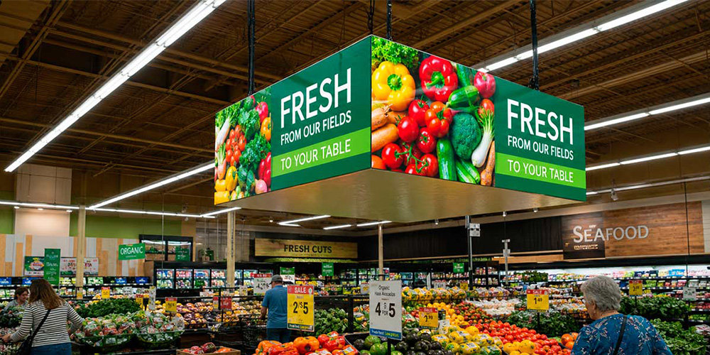 Grocery store produce section with hanging green banners reading 'Fresh from our fields to your table' over colorful vegetables and shoppers browsing.