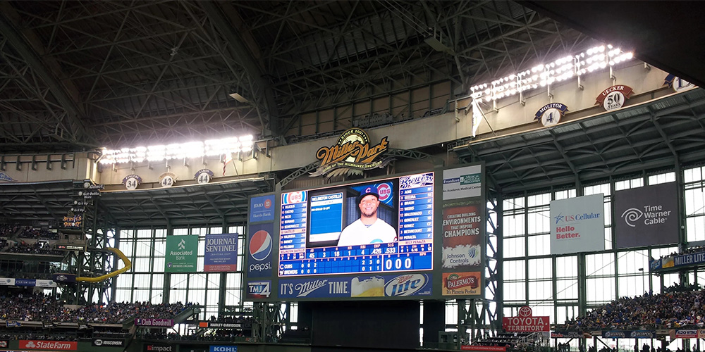 Interior of a baseball stadium with a large central scoreboard displaying a player, surrounded by advertisements and seating sections.