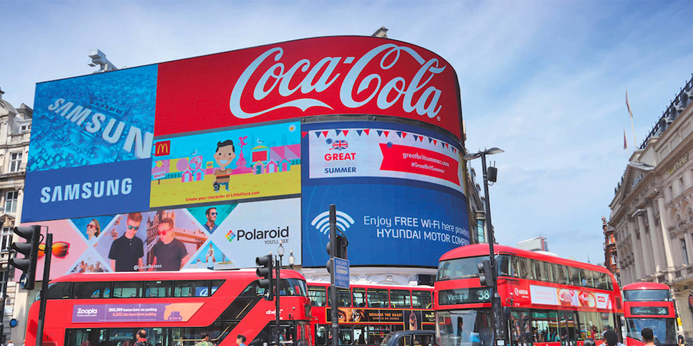 Billboard at a busy city square featuring Coca-Cola and Samsung ads with red double-decker buses in front, London-style scenery.