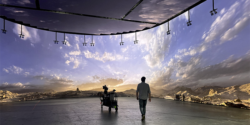 卷曲屏幕 - LED Screen Board - LED Screen Board Person walks away on a movie set at sunset, with a camera dolly and overhead lights, desert mountains in the distance.