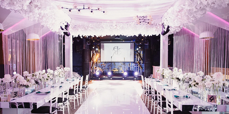 室内婚礼屏幕3 - LED Screen Board - LED Screen Board Wedding reception hall with long white tables, white floral centerpieces, pink/purple uplighting, and a stage with projection screen.