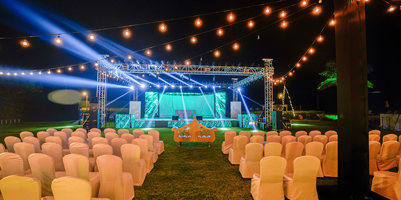 室外婚礼屏幕1 - LED Screen Board - LED Screen Board Outdoor night event setup: rows of white chairs facing a lit stage with string lights overhead and blue spotlighting on stage.