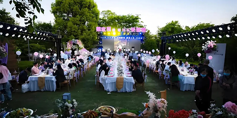 室外屏幕2 - LED Screen Board - LED Screen Board Outdoor banquet scene at dusk with long rows of tables, guests dining under string lights and pink decor, a stage at the far end with a screen.