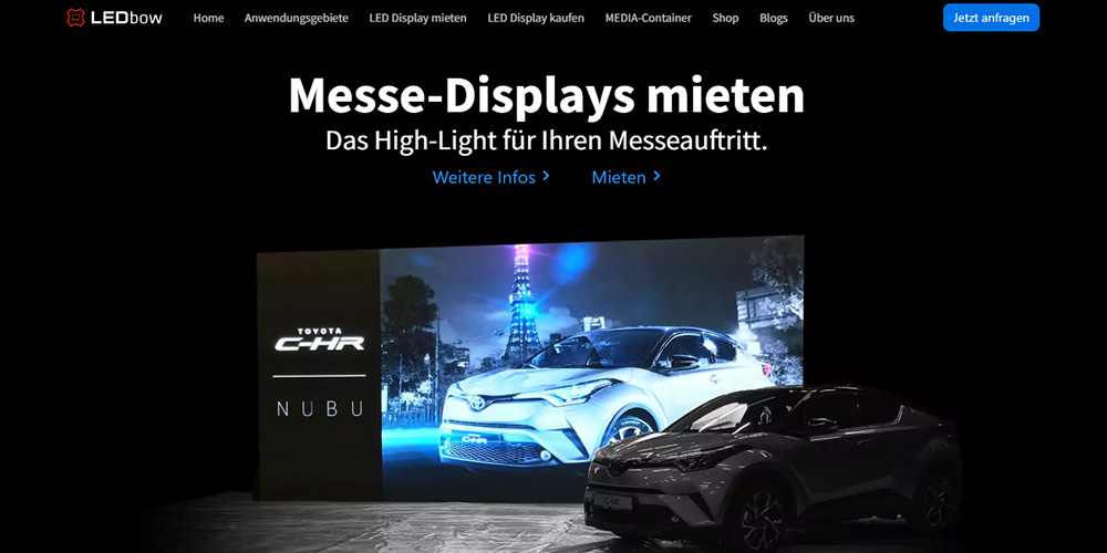 Großes LED-Display auf einer Messe, das ein blaues Toyota C-HR-Visual zeigt; daneben ein dunkles Auto auf der Bühne im Hintergrund.