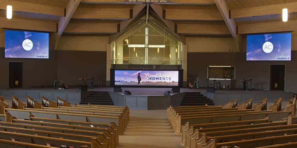 教堂LED - LED Screen Board - LED Screen Board Wide view of an empty auditorium with wooden pews, a raised stage, and three screens displaying 'Moments' and AC logos.