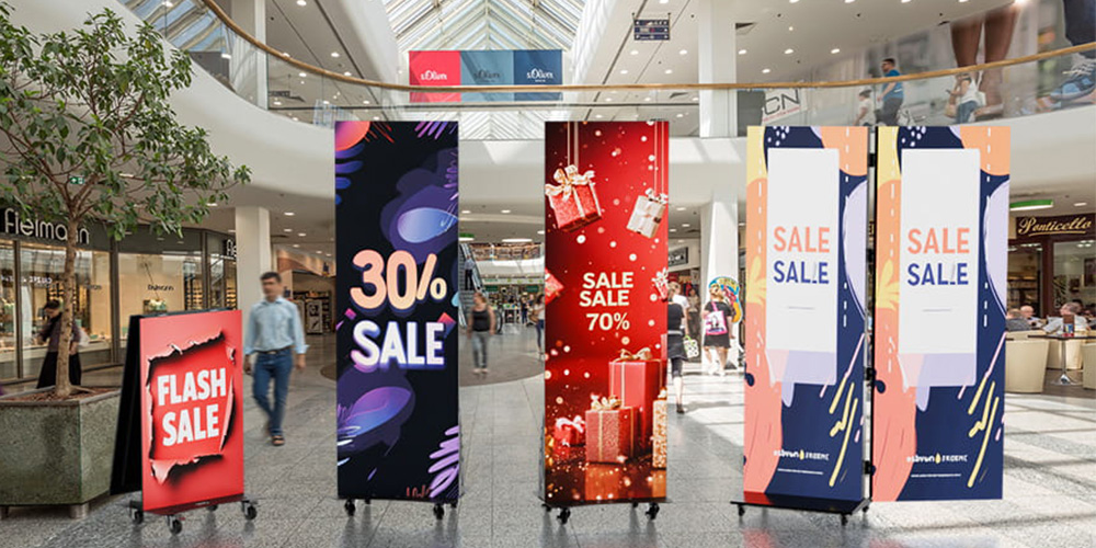 海报屏幕6 - LED Screen Board - LED Screen Board Indoor mall with tall sale banners: 30% SALE, red gift box banner, and two 'SALE' signs in bright colors near shoppers.