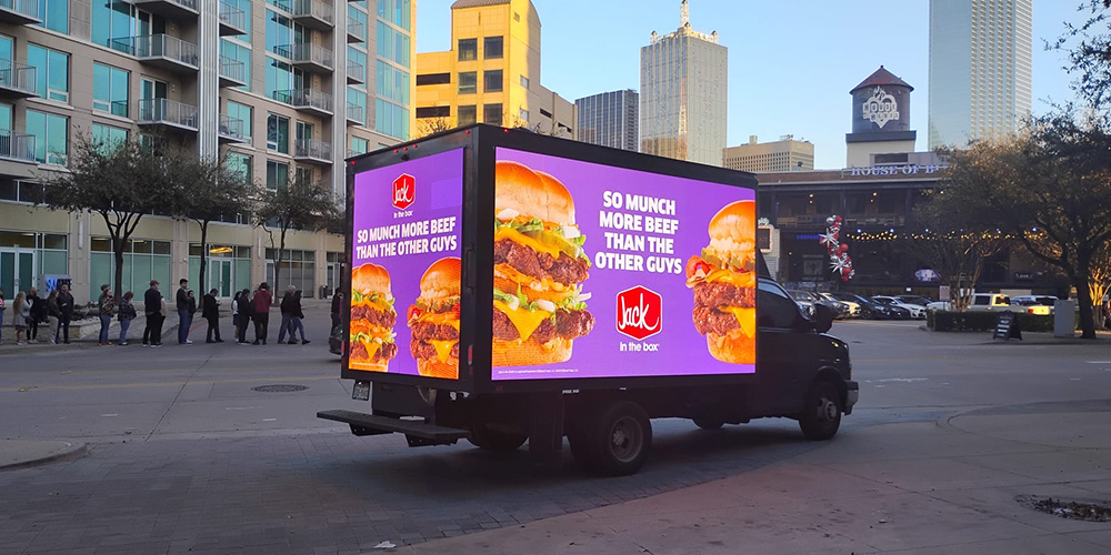 01-truck-billboard-advertising-city-street - LED Screen Board - LED Screen Board Box truck with a bright purple burger advertisement on its side in a city square; pedestrians line up nearby.