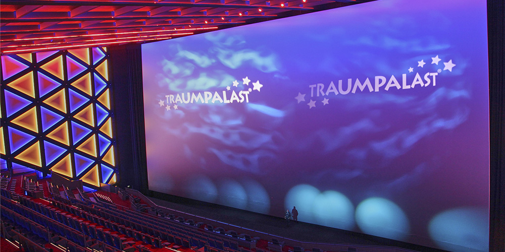 04-Traumpalast-Leonberg - LED Screen Board - LED Screen Board Wide theater interior with a giant screen showing TRAUMPALAST logos and star graphics, red seats and geometric wall panels on the left.