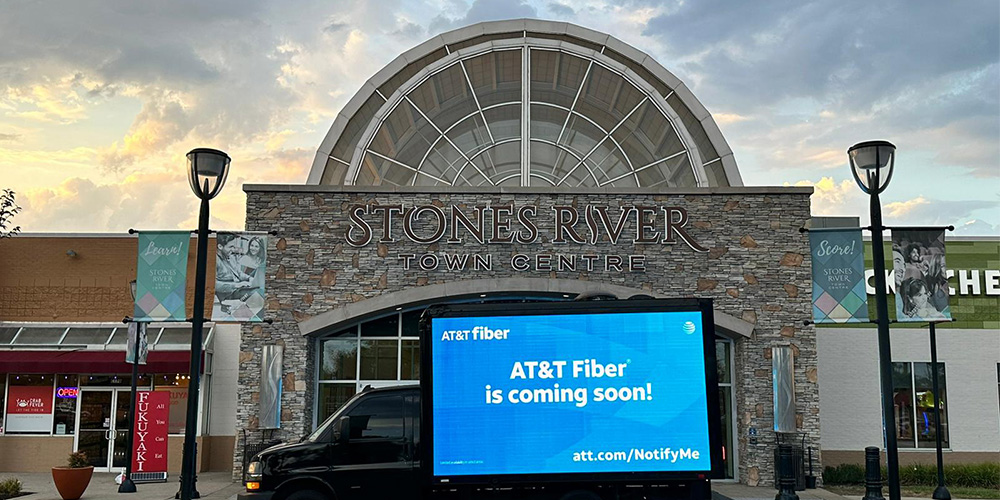 04-mobile-led-billboard-truck-shopping-center - LED Screen Board - LED Screen Board Stone River Town Centre mall with a large blue AT&T Fiber screen on a truck in front of the entrance, sunset sky above.