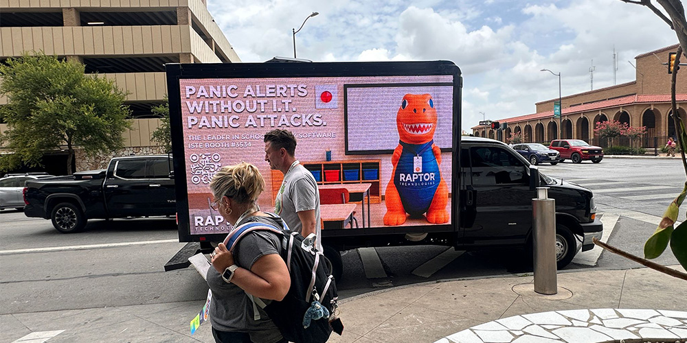 06-truck-billboard-advertising-urban-pedestrians - LED Screen Board - LED Screen Board Mobile billboard truck on a city street displaying a dinosaur mascot and the text 'Panic Alerts Without It' while pedestrians walk by.