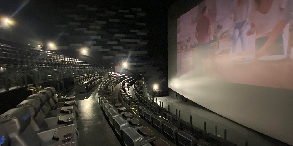 10-Royal-Cinemas-IMAX-Pooler - LED Screen Board - LED Screen Board Empty cinema auditorium with curved rows of seats facing a large screen, dim lighting and a blurred image on screen.
