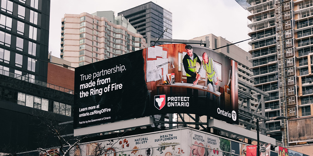 Large curved billboard in a city promoting Ontario's 'Protect Ontario' campaign, showing workers and the text 'True partnership, made from the Ring of Fire'.