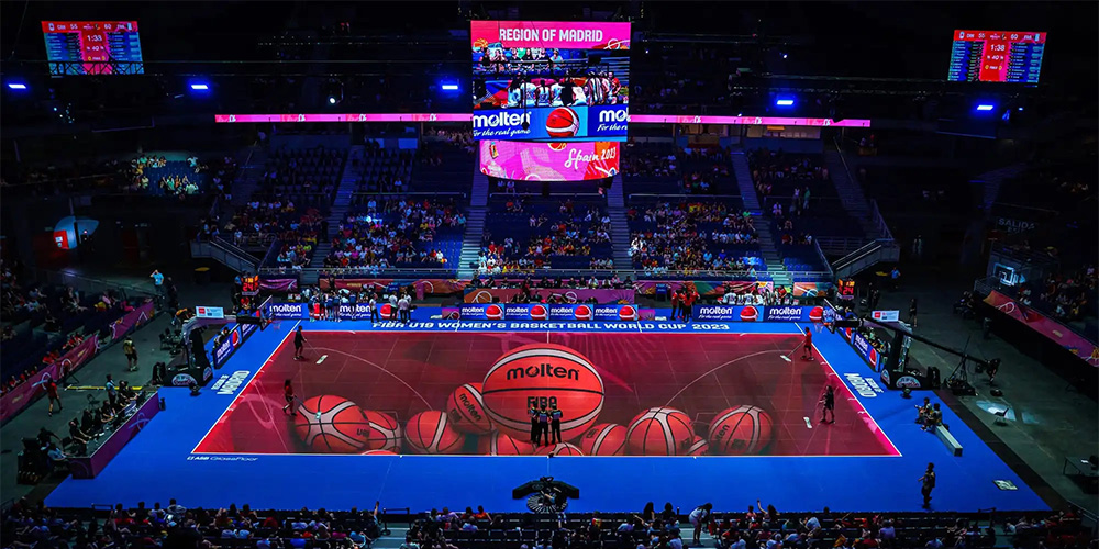 Indoor basketball arena filled with spectators watching a game on a red Molten-branded court with oversized ball graphics and bright blue sidelines