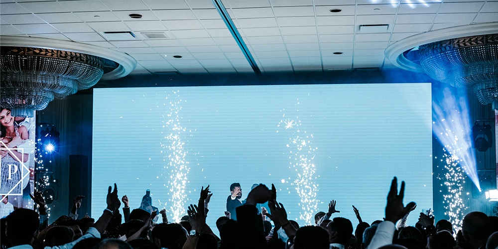 dimension led1 - LED Screen Board - LED Screen Board Crowd raises hands at an indoor concert with a DJ on stage and pyrotechnic sparks near a large blue LED screen.