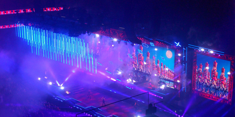 Concert stage at night with blue neon lights and large screens showing performers, surrounded by purple lighting and an excited audience in the dark arena