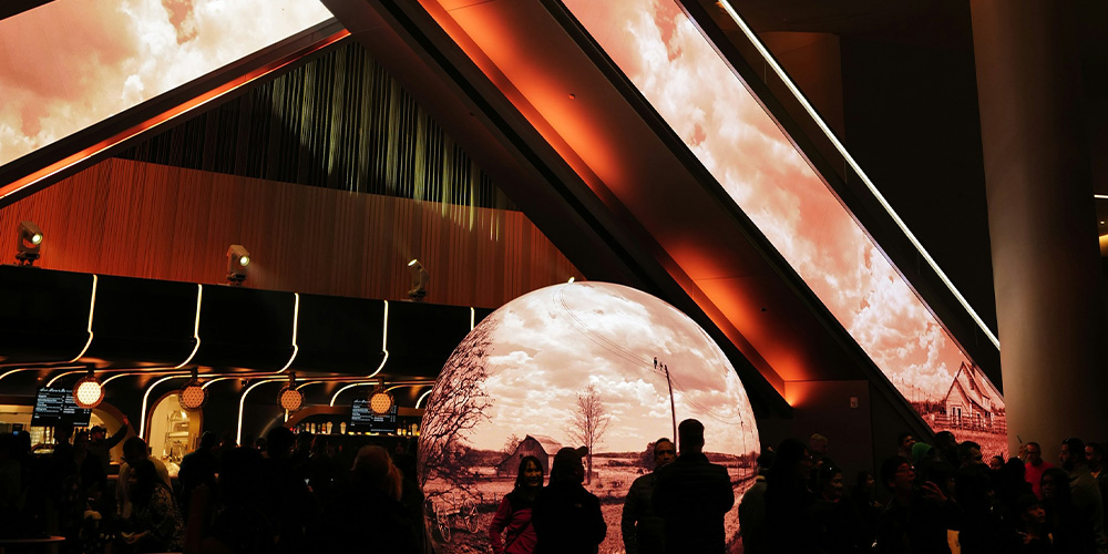 dimension led3 - LED Screen Board - LED Screen Board Crowd of visitors gathers around a large reflective globe showing a rural landscape in an orange-lit, modern gallery space.