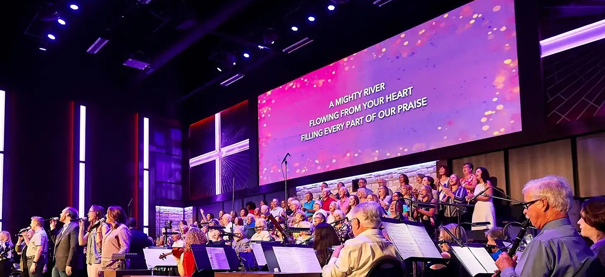 Church-LED-Screen-7 - LED Screen Board A large choir and musicians perform on a connected stage under purple lights, with a screens displaying lyrics above them.