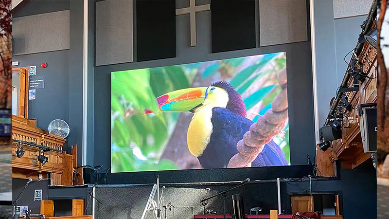 Church-LED-Screen-8 - LED Screen Board Large indoor stage with a big screen displaying a colorful toucan, a dark wall with a cross symbol above, and stage equipment nearby.