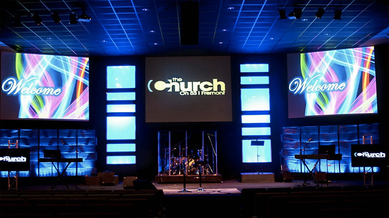 Church-LED-Screen-9 - LED Screen Board Stage in a church auditorium with colorful Welcome screens on both sides, a central screen showing the church name, and a drum kit and keyboards set up for worship.