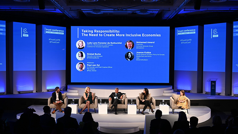 Event-LED-Screen-7 - LED Screen Board Panel of five speakers on a blue-lit conference stage, with a large screen displaying the talk title behind them and an audience in the foreground.