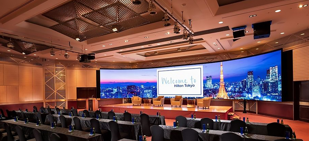 Hotel-LED-Display - LED Screen Board Large conference room with a curved LED screen on stage displaying 'Welcome to Hilton Tokyo' and several chairs for speakers in front of the audience seating area.