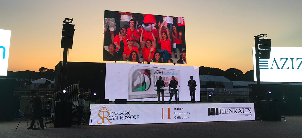 Mobile-LED-Screen-6 - LED Screen Board Outdoor stage at sunset with a large screen showing cheering fans; three presenters on stage and sponsor banners along the front.