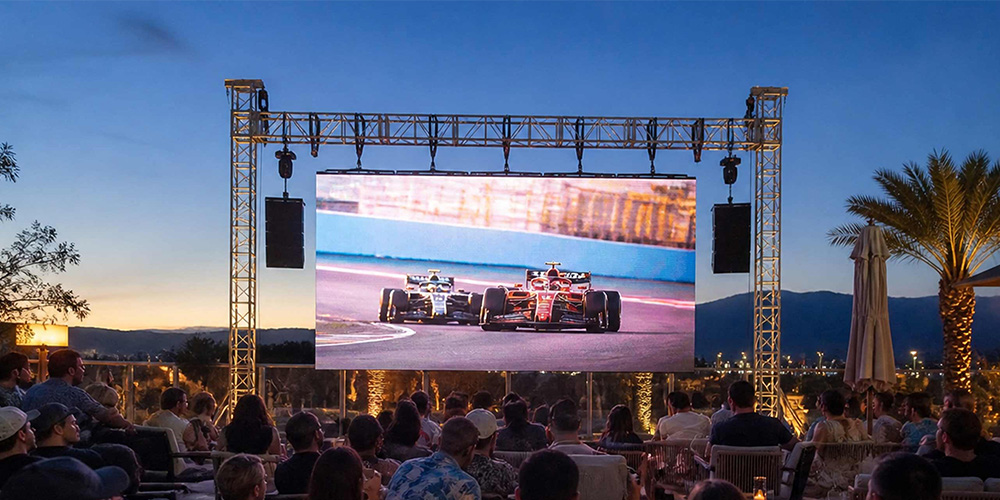 waterproof-led-display-outdoor-event - LED Screen Board Outdoor audience watching a large screen showing a Formula 1 race at sunset, with metal scaffold stage and palm trees nearby